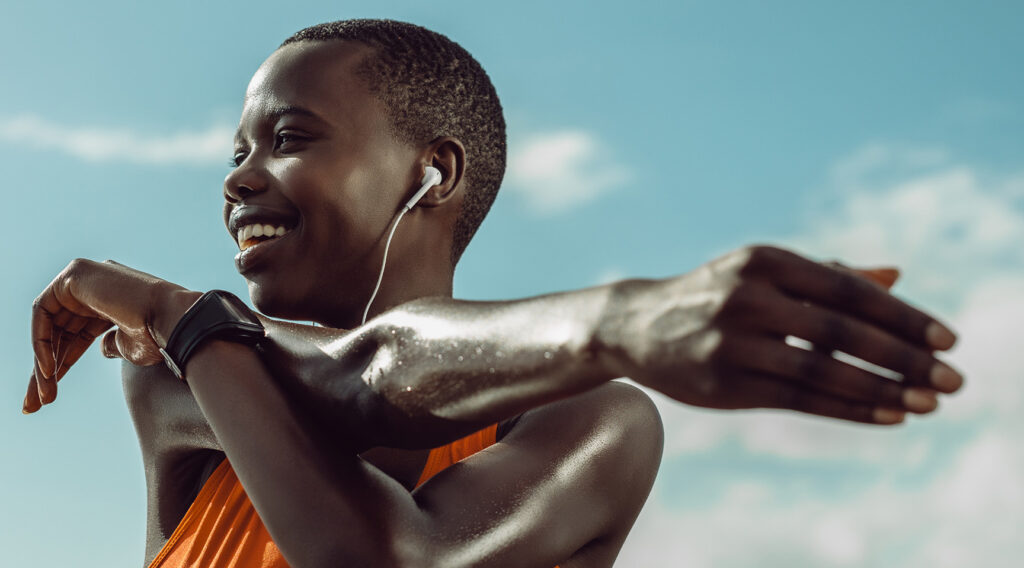 Female, listening to music through white earphones and wearing an orange top, is warming down after a run in the sun.