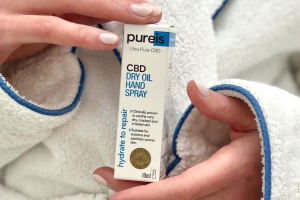 Close up of the front of a CBD Dry Oil Hand Spray Carton. Held by a person wearing a white bathroom robe.